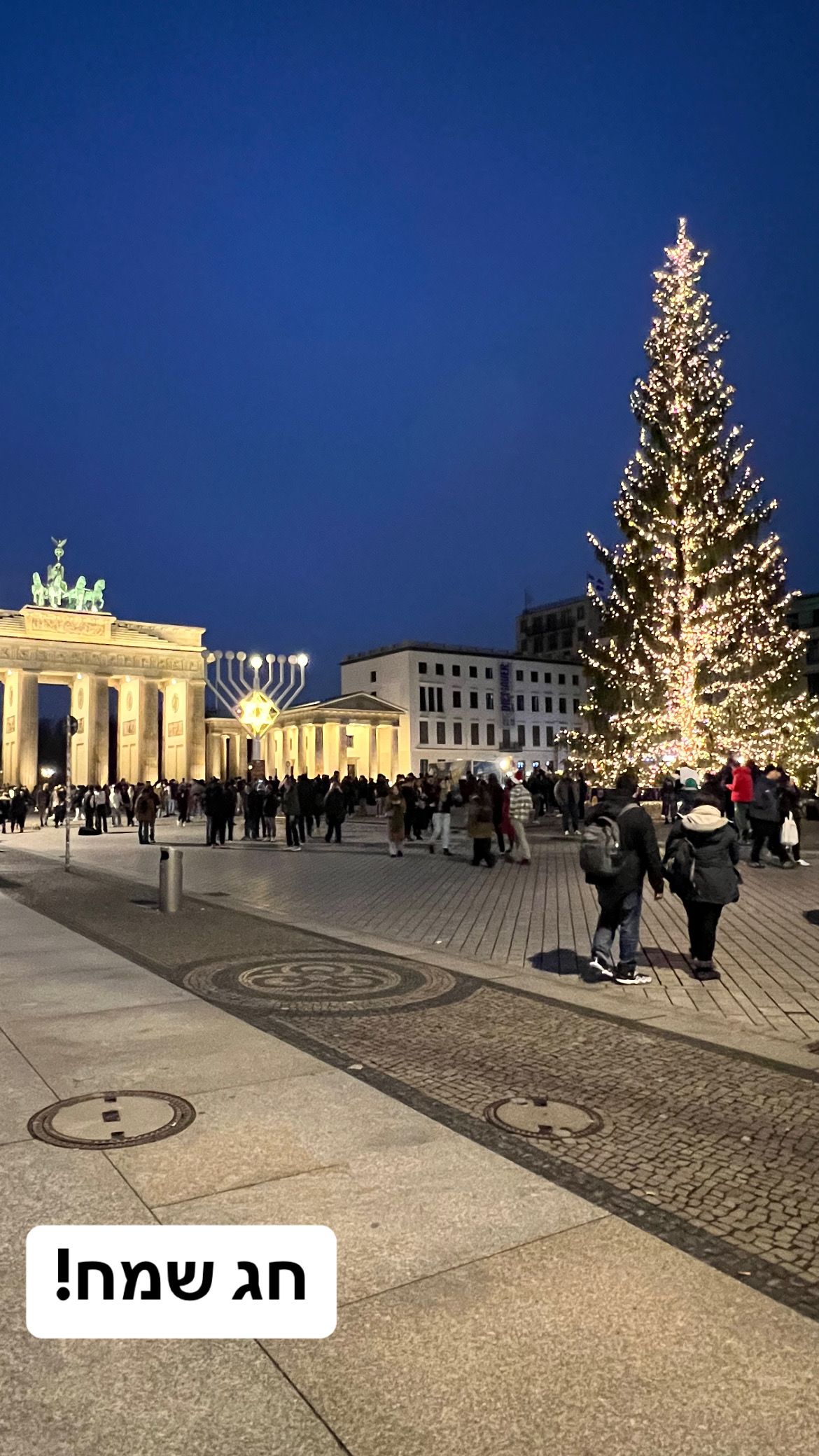שער ברנדנבורג עם החנוכיה ועץ חג המולד העצום בשעת ערב. עדיין מוצף באנשים. כיתוב: חג שמח!